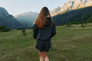 Naklejka premium A solitary woman stands in a grassy alpine meadow, facing distant mountains as sunlight warms the scene, creating a peaceful outdoor moment, with quiet skies and expansive horizon.