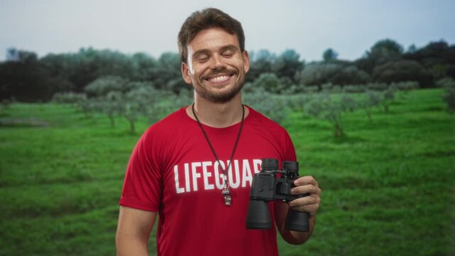 Young hispanic man lifeguard holding binoculars and whistle necklace, smiling with eyes closed and flashing peace sign in forest; calm vigilance.