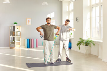 Professional physiotherapist instructing male patient on post-traumatic exercises using resistance band with handles. Hands muscles and joints recovery at rehabilitation center concept.