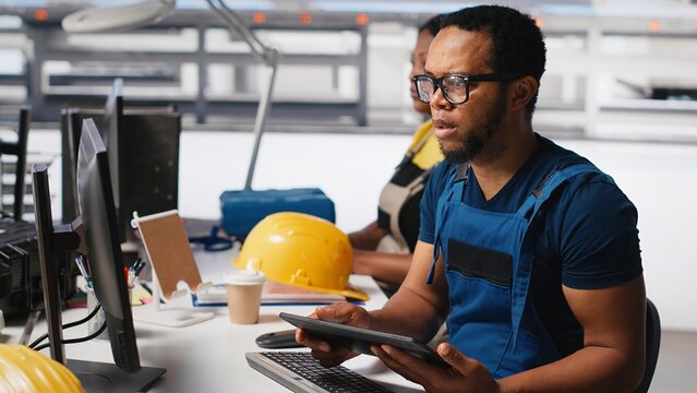 Engineer investigating malfunctioning systems in photovoltaics factory, comparing parameters with pc and tablet. Black man inspecting solar panels manufacturing plant, high tech. Camera A.
