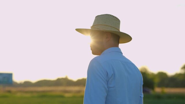 Close up portrait of handsome male in aht standing in countryside at sunset or sunrise and looking away. Young man farmer in green field alone. Agricultural activity, farmland concept