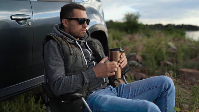 A man is resting on a car trip on the coast of a lake, ocean or sea. He has a seat in a folding chair. He is holding a thermos or thermo mug
