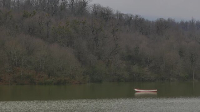 A single white and red boat floats silently on a calm lake, surrounded by dense leafless forest in winter. The muted tones and still water create a serene, meditative atmosphere of solitude and reflec