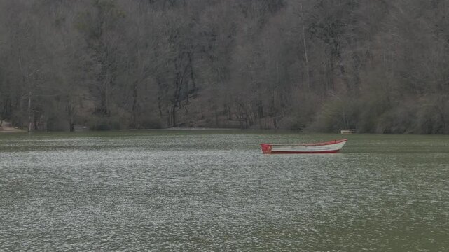 A single white and red boat floats silently on a calm lake, surrounded by dense leafless forest in winter. The muted tones and still water create a serene, meditative atmosphere of solitude and reflec