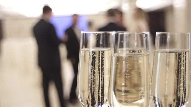 Champagne bellows rise in glasses at party. Foreground glasses with drink, background people talk. Close-up, slow-motion, concept.