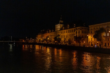 Fototapeta premium Illuminated historic baroque building with clock tower on riverside embankment at night with golden reflections in dark water. Concept of urban nightscape, architectural heritage, romantic atmosphere.