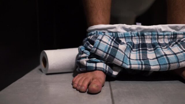 Video process of man's hand reaching to toilet paper roll after defecation and cut it. Guy sit on pot in rest room with pants on his feet. Alone in wc or toilet. Close up.