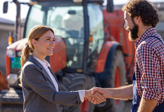 Farmer and businesswoman shaking hands in front of tractor
