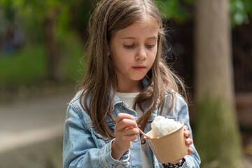Young girl in denim jacket enjoying cup of ice cream in park. Cute child is enjoying frozen desserts in park against blurred trees on sunny summer day 