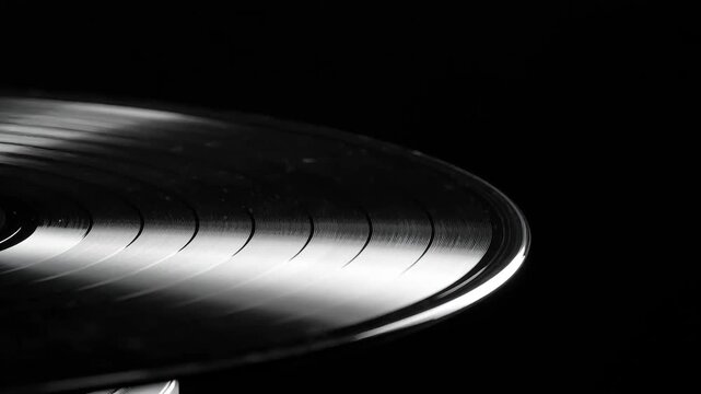 Extreme close-up of a black vinyl record spinning on a turntable, with groove patterns reflecting light, on a dark, black background