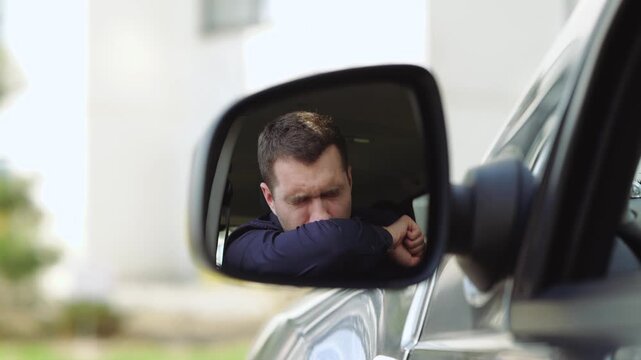 Young man inside car. A view from side mirror of businessman caughing hard and deep. Cover mouth with hand. Alone in car. Blurred background.