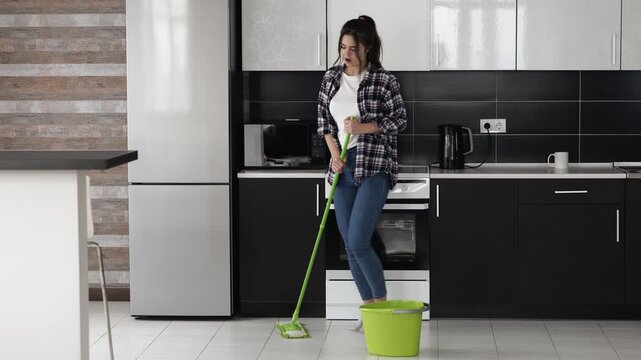 Young woman in kitchen during quarantine. Wiping floor with mop and dance. Singing song during cleaning. Enjoy her time in apartment.