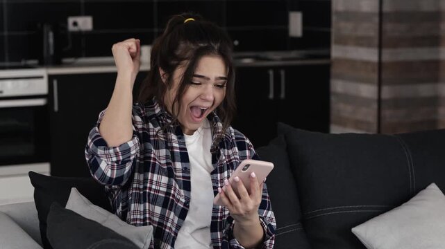 Young woman in kitchen during quarantine. Sit on sofa and using smartphone. Start to scream with joy and happiness. Cheerful positive person.