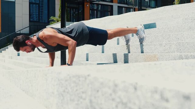 Young man exercising outside. Sporty guy doing push up exercises on steps on street. Fitness activity, exercising or training alone. Listen to music in earphones.