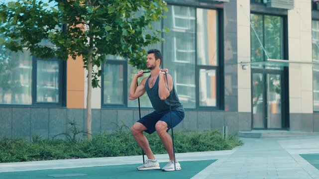 Young man exercising outside. Video camera moving backwards. Slow motion of sportsman holding rubber band and training. Doing squat exercise. Muscled powerful guy training on street.