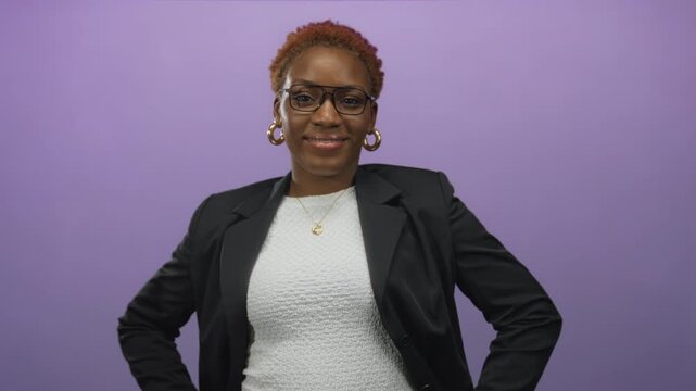 Woman wearing glasses and a black blazer, smiling while standing with hands on hips in a purple studio; confidence empowerment strength poise.