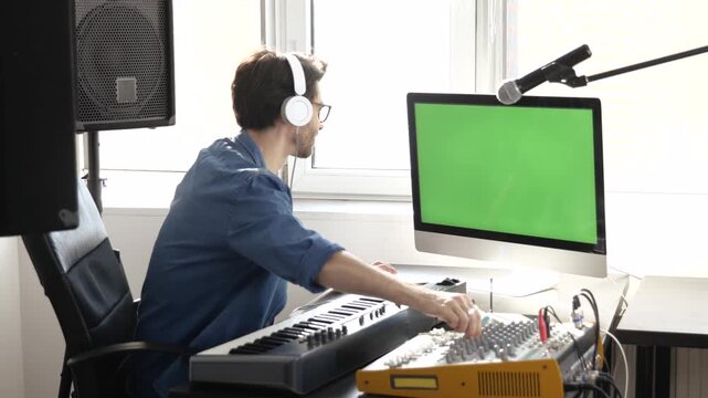 Young man in sound recording studio. Using and adjusting console mixer and enjoying listening to music in headphones. Big green screen.