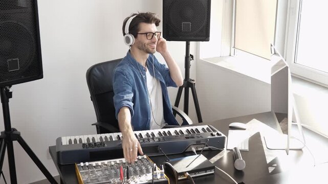 Young man in sound recording studio. Sit in front keyboard and adjusting sound mixing console. Listening to music in headphones.