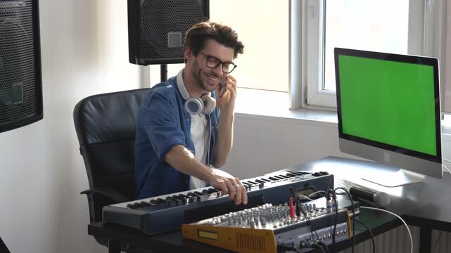 Young man in sound recording studio. Guy sit in front keyboard and talking on phone. Adjusting music console during phone conversation.