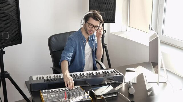 Young man in sound recording studio. Sit at table and using mixing console. Listening to music through headphones.