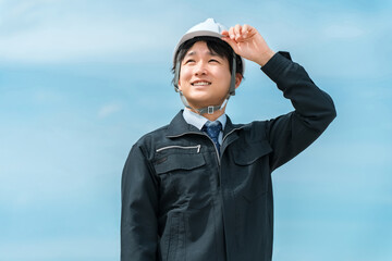 青空の下にいる作業着姿のヘルメットを被った男性
