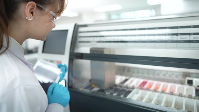 A histotechnologist operates an automated stainer, using a tablet to manage the protocol as slide racks with tissue samples move through a series of reagent baths for Hematoxylin and Eosin staining.