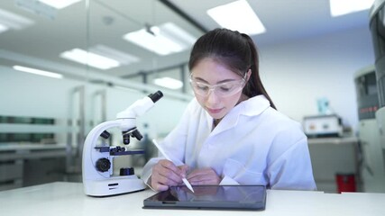 A microbiologist records research data on a digital tablet. She is conducting pharmaceutical analysis or pathology testing in a high tech biotech lab with a microscope. - Powered by Adobe