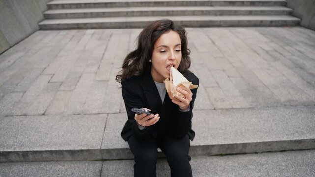 Caucasian girl with brown long hair sitting on stairs in downtown eating delicious sandwich, scrolling her smartphone.