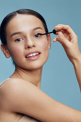 Fototapeta premium Young woman applying mascara with a bright smile, showcasing beauty and confidence against a soft blue backdrop, emphasizing makeup and self care
