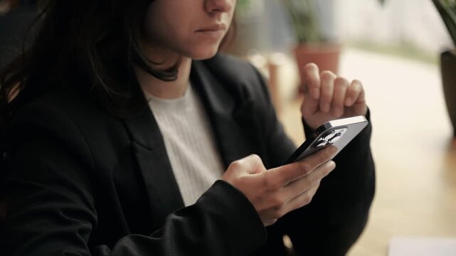Pretty Caucasian woman with dark long hair sitting in a modern restaurant, scrolling, and typing on her smartphone. Girl is creating content, and loos serious and tired.