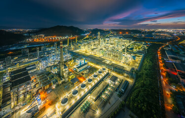 Fototapeta premium Panoramic of a huge industrial complex and petrochemical plant illuminated at night surrounded by green hills and roads.