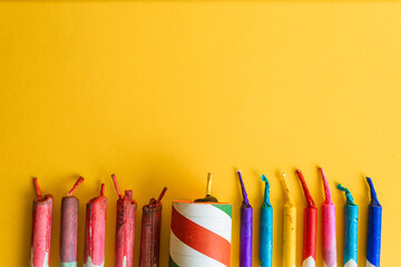 A line of bright, colorful firecrackers and a large striped explosive, leaving ample blank space on a vibrant yellow background.
