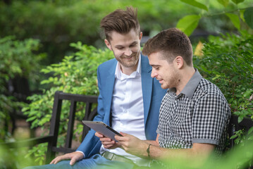 Two businessman or gay friend partner sitting at chair in park while using digital tablet and paper work plan for talk discussion sharing analyze data together while consulting.