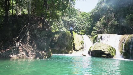 Slow motion of a Southeast Asian man in mid-air jumping off a rocky cliff into the clear turquoise river pool near a beautiful tropical waterfall.
 - Powered by Adobe
