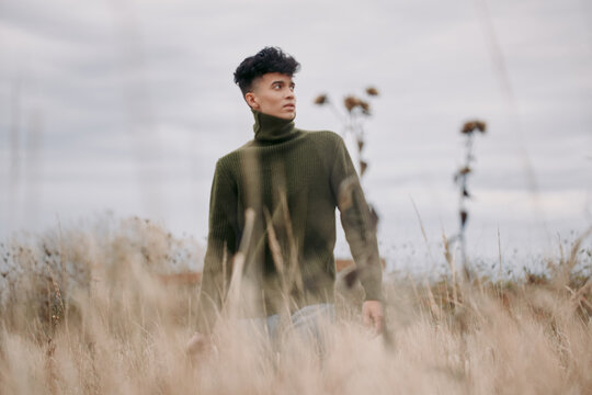 A young man wearing an olive turtleneck stands in tall field grass, captured in an authentic, credible portrait that emphasizes natural texture, muted tones and a thoughtful expression.