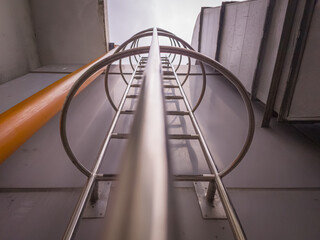 Upward view of a vertical metal ladder with safety hoops in an industrial setting. Strong geometric lines and perspective highlight height, structure, and safety design.
