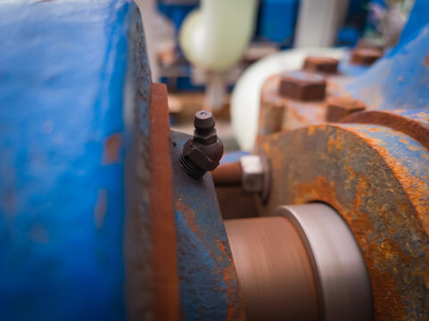 Close-up of a grease fitting (zerk) on rotating industrial machinery. Visible rust, bolts, and motion blur highlight active lubrication in a heavy-duty environment. - Powered by Adobe