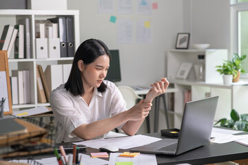 Young Asian Businesswoman suffering from wrist pain while working on computer, Businesswoman massaging her hand and wrist after long hours in front of a computer, Healthcare office syndrome concept