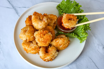 A close-up shot of a golden brown fried shrimp being dipped into a small glass bowl of red dipping sauce. Served on a white plate with fresh green lettuce in the background.