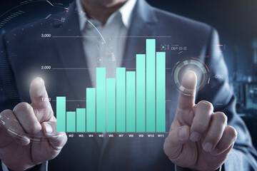 A close-up image of a businessman gesturing towards an expanding virtual screen displaying a business performance bar graph.
