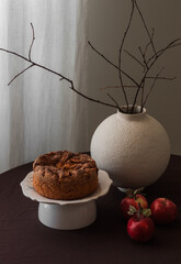 A pie, apples, a vase with decorations on a table with a brown tablecloth in the living room