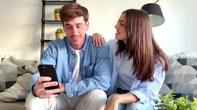 Happy couple sitting together on sofa at home, using smartphone and smiling while spending time together, concept of love, relaxation, communication, modern lifestyle and positive relationship