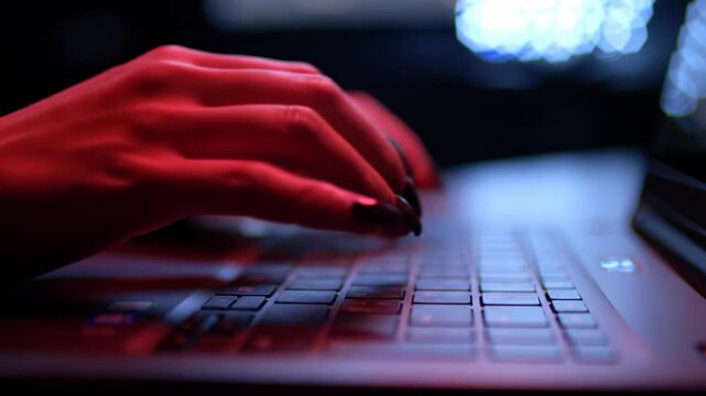 Elegant hands with glossy dark nails frantically type on a sleek, dark laptop keyboard, intensely lit by dramatic crimson red light against a blurred high-tech environment. Urgent cybersecurity task
