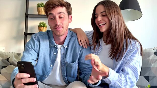Happy couple sitting together on sofa at home, using smartphone and smiling while spending time together, concept of love, relaxation, communication, modern lifestyle and positive relationship