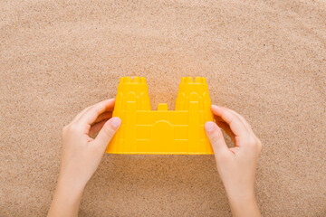 Baby hand holding and showing yellow plastic castle tower shape on light brown sea sand background. Little child activities at sea beach. Closeup. Point of view shot. Top down view.