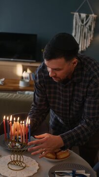 Vertical shot of young Jewish man placing shammash after lighting up all nine candles on hanukkiah, celebrating last day of Hanukkah in cozy modern apartment