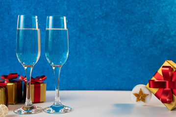 Two champagne glasses with Christmas presents and decorations on a blue background. Perfect for New Year and holiday celebrations. 