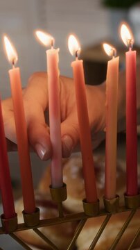Vertical cropped slowmo of hands of unrecognizable woman putting burning shammash on hanukkiah, preparing festive table for Hanukkah celebration at home