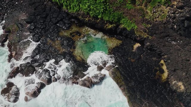 Aerial top-down view of the rugged volcanic coastline at Roche Qui Pleure, Mauritius, with waves crashing against dark rocks and a turquoise tide pool in a dramatic ocean landscape.