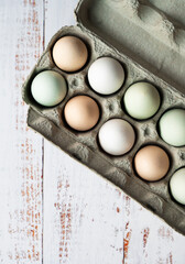 Top view of carton of colorful eggs on light wood background.